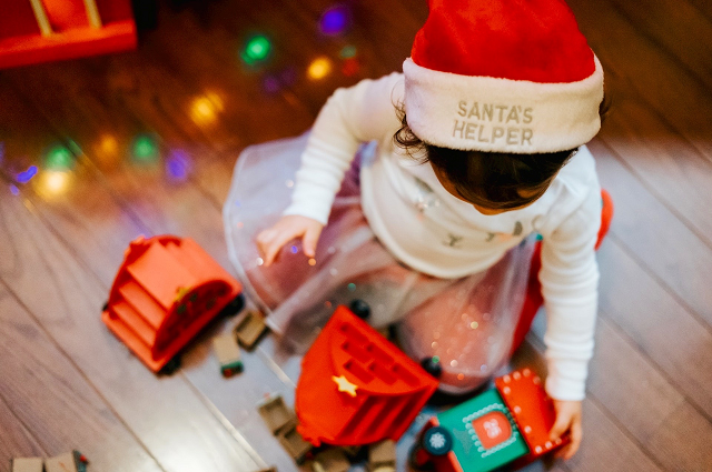 25 a menos de 25 dólares: Los mejores regalos de Navidad para tu bebé, a baby girl wearing a Santa hat and playing on the floor with toys with holiday lights reflected on the wooden floor.